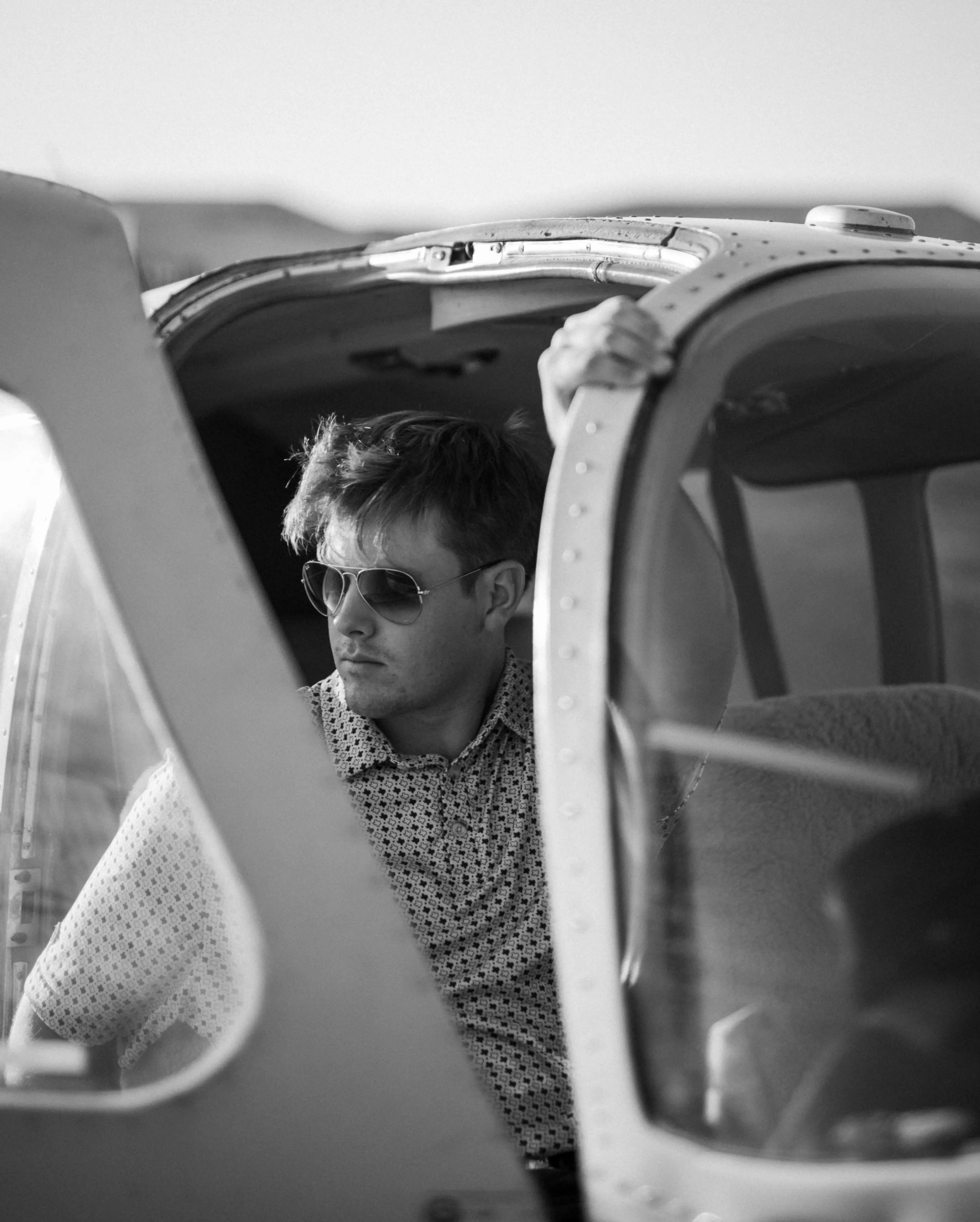 Black-and-white editorial portrait of a man in sunglasses leaning out of a vintage airplane cockpit