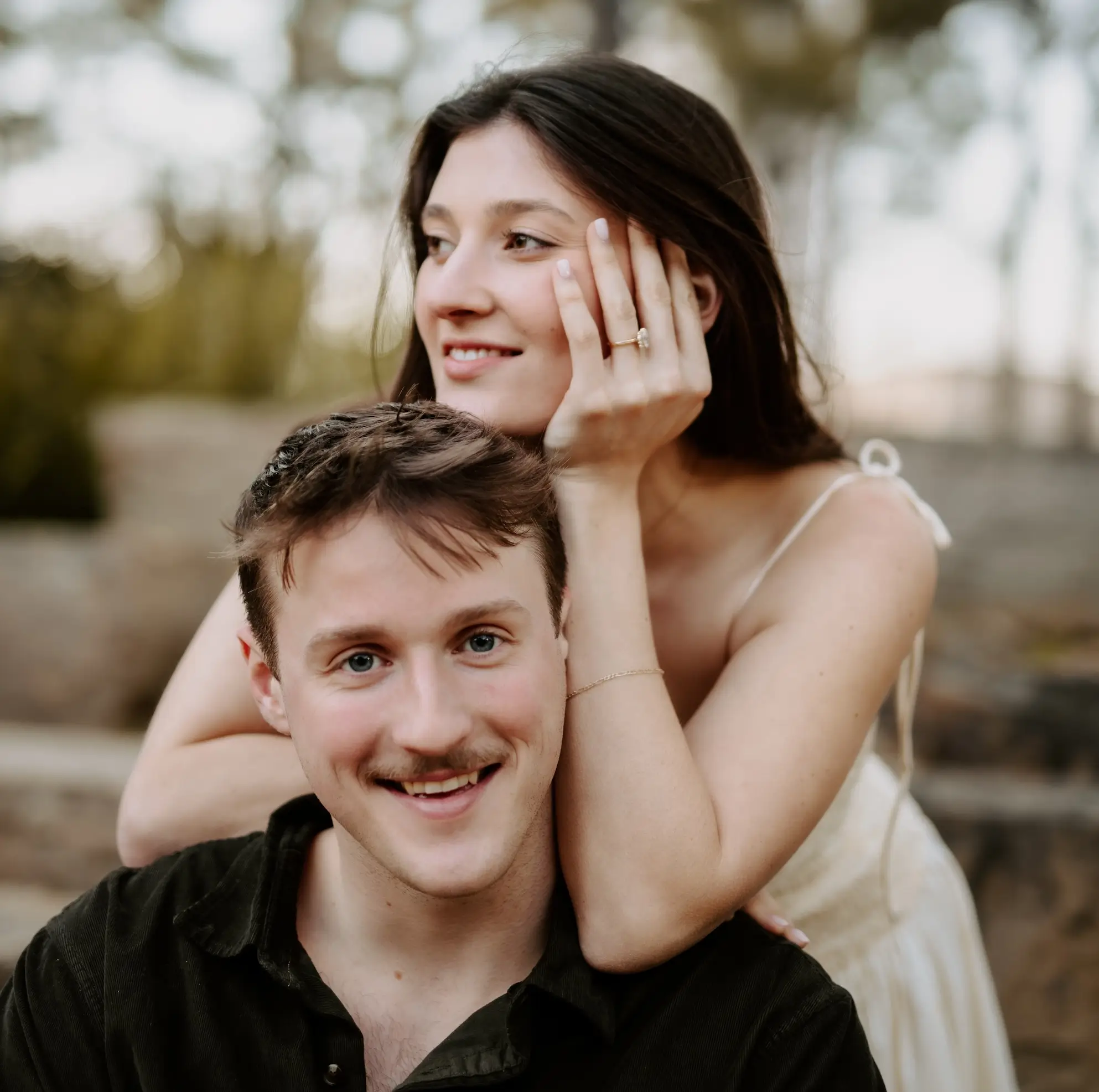 Newly engaged couple smiling together outdoors, her engagement ring visible as she rests her hand near his head
